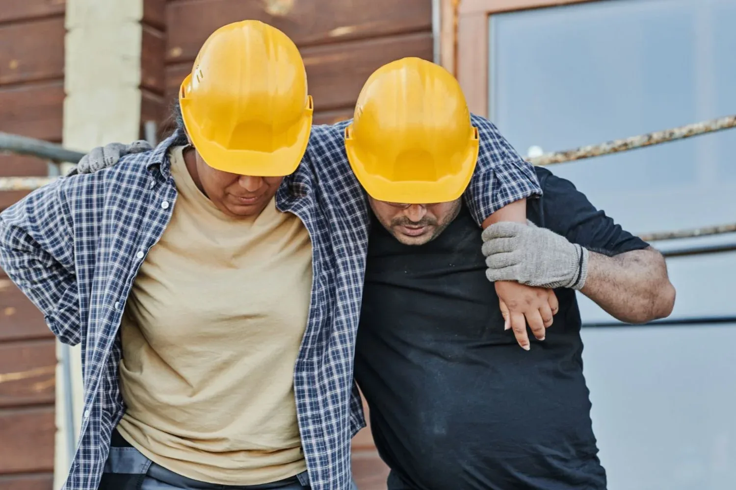 Two construction workers in hard hats, one supporting the other who is limping